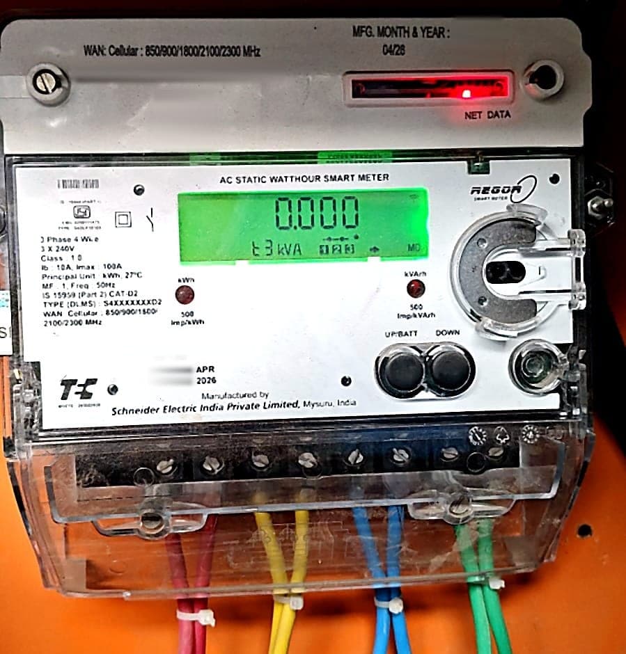 A three-phase DLMS smart meter installed on an electrical panel, its green display lit, with red, yellow, blue, and green cables wired into the terminal block.